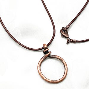 May include: A copper-colored pendant necklace with a circular design. The pendant is attached to a brown leather cord with a clasp. The necklace is displayed on a white surface.