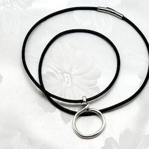May include: A minimalist necklace featuring a black cord and a silver-toned pendant. The pendant is a simple, open circle design. The necklace has a magnetic clasp. The jewelry is displayed on a white floral background.