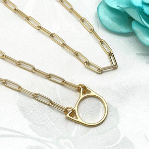 May include: Gold-colored chain necklace with a cat-shaped pendant. The necklace features a delicate paperclip chain design. The pendant is a stylized cat face. The jewelry is displayed on a white surface with a blue flower in the background.