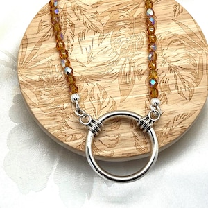 May include: A silver-toned pendant necklace featuring a large, open circle. The necklace has a strand of amber-colored, faceted beads. The beads are connected to the circle pendant with silver-toned findings. The necklace is displayed on a wooden surface.