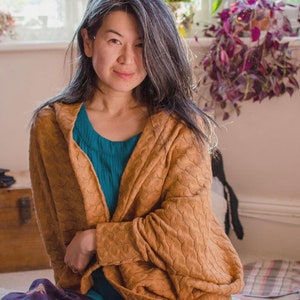 May include: A woman with long gray hair is wearing a brown knitted cardigan over a teal blue shirt. She is sitting on a patterned cushion with a purple and black design.