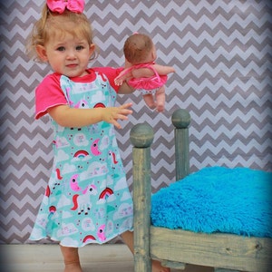 May include: A young girl in a pink and blue dress with a unicorn and rainbow print holds a doll in a pink dress. The girl is standing next to a small wooden bed with a blue fuzzy mattress.