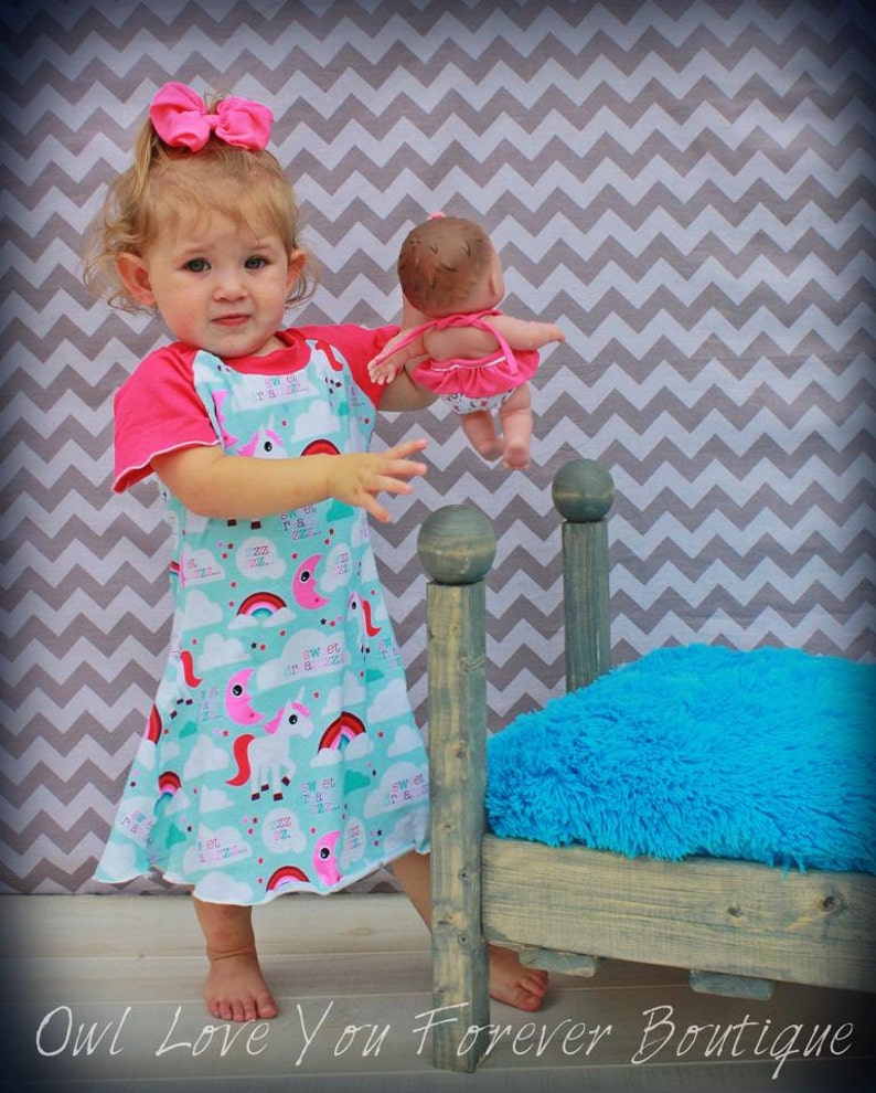 May include: A young girl in a pink and blue dress with a unicorn and rainbow print holds a doll in a pink dress. The girl is standing next to a small wooden bed with a blue fuzzy mattress.