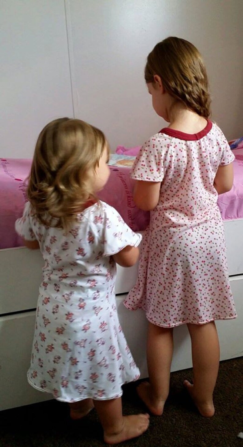 May include: Two young girls wearing white and pink floral print nightgowns. The girl in the front is looking at a book. The girl in the back is looking at the girl in the front. The girls are standing in front of a white dresser with a pink and white bedspread.
