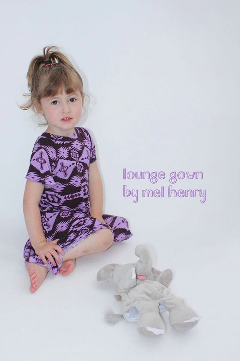 May include: A young girl wearing a purple and brown patterned lounge gown sits on a white background with a gray elephant stuffed animal. The text "lounge gown by mel henry" is written in purple on the white background.