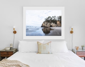 May include: A framed photograph of a rocky cliff with trees on top, overlooking a beach with calm water reflecting the sky. The photograph is hanging above a white bed with a white pillow and a patterned throw pillow.