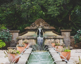 May include: A stone fountain with a cascading waterfall in a garden setting. The fountain is surrounded by lush greenery and potted plants.