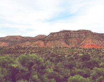 May include: A scenic landscape featuring a mountain range with red and brown rock formations. Lush green trees and shrubs fill the foreground, with a cloudy blue sky above. The image captures a natural, outdoor setting.
