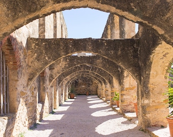 May include: A long, narrow hallway with a series of stone arches. The arches are made of rough-hewn stone and are supported by pillars. The hallway is lit by sunlight streaming in from the top of the arches. The floor is made of gravel.