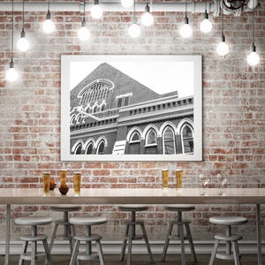May include: A black and white framed print of a building with arched windows hangs on a brick wall. Below, a long wooden table is set with stools and glasses. Overhead, Edison-style light bulbs hang from the ceiling.