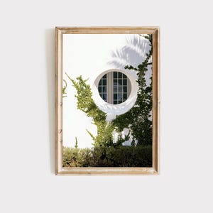 May include: A framed print featuring a white wall with a circular window. Green vines and foliage climb the wall, framing the window. The print has a light brown wooden frame, and the image is well-lit.