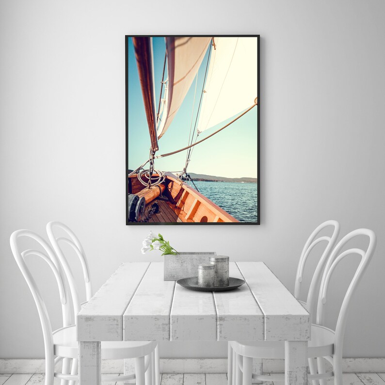 May include: A close-up view from the deck of a sailboat, showing the wooden deck and the white sails billowing in the wind. The blue water and a distant shoreline are visible in the background.