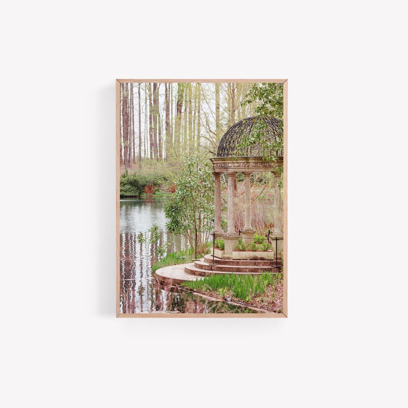 May include: A stone gazebo with a black metal roof sits on a small island in a pond surrounded by trees. The gazebo has steps leading up to it and is surrounded by lush greenery.