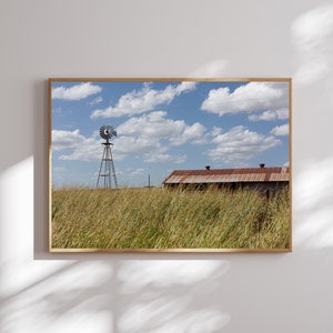 May include: A vintage windmill stands tall in a field of tall grass, with a rustic barn in the background. The sky is blue with fluffy white clouds.