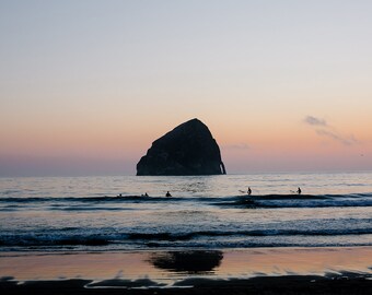 May include: Silhouette of a large rock formation in the ocean at sunset with people surfing in the foreground.