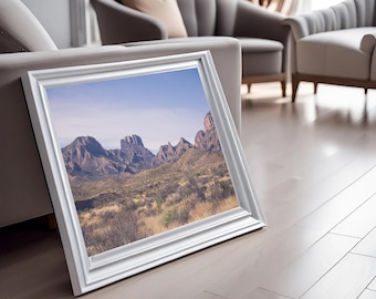 May include: A framed photograph of a mountain range with a desert landscape in the foreground. The mountains are a light brown color and the sky is a light blue. The frame is white.