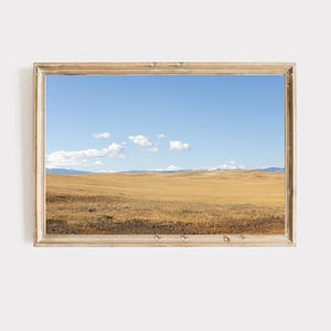 May include: A framed landscape print featuring a vast, golden field under a bright blue sky with scattered white clouds. The artwork is enclosed in a light-colored wooden frame, adding a rustic touch to the serene scene.