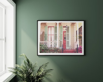 May include: A vintage photograph of a house with a red door and green shutters. The house has a white porch with a railing and a small yard with green plants.