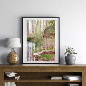 May include: A stone gazebo with a metal roof sits on a small island in a pond surrounded by trees. The gazebo has a stone path leading up to it and is surrounded by green grass and trees.