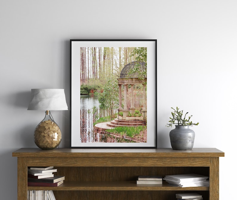 May include: A stone gazebo with a metal roof sits on a small island in a pond surrounded by trees. The gazebo has a stone path leading up to it and is surrounded by green grass and trees.