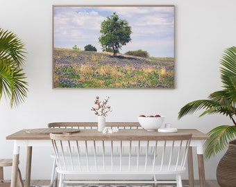 May include: A framed landscape print featuring a vibrant field of purple wildflowers and a lone tree under a cloudy sky. The print is displayed above a wooden dining table with white chairs, a patterned rug, and potted plants.