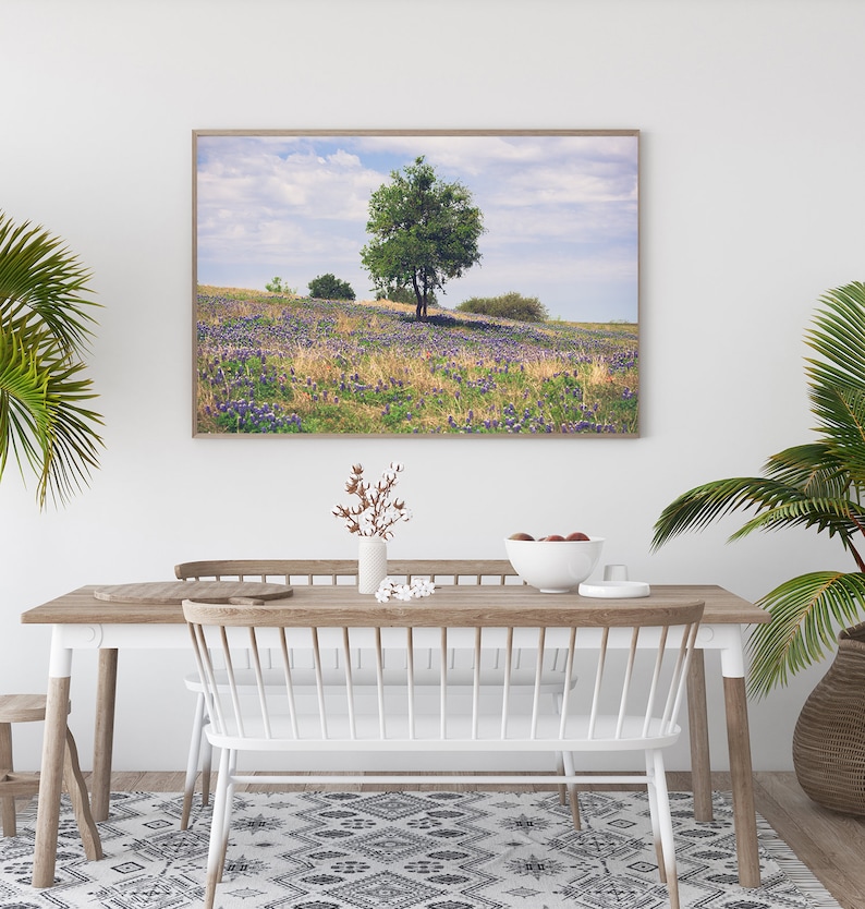 Op de afbeelding: Een ingelijste landschapsafdruk met een veld vol paarse wilde bloemen en een eenzame boom onder een bewolkte hemel. De afdruk hangt boven een houten eettafel met witte stoelen, een patroonkleed en potplanten.