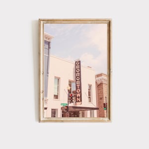 May include: A framed photograph of a building with a sign that reads "GEORGETOWN" in a vintage style. The building is a light pink color with white trim and has a red and green traffic light in front of it.