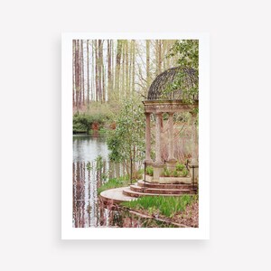 May include: A stone gazebo with a black metal roof sits on a small island in a pond. The gazebo has steps leading up to it and is surrounded by green trees and bushes.
