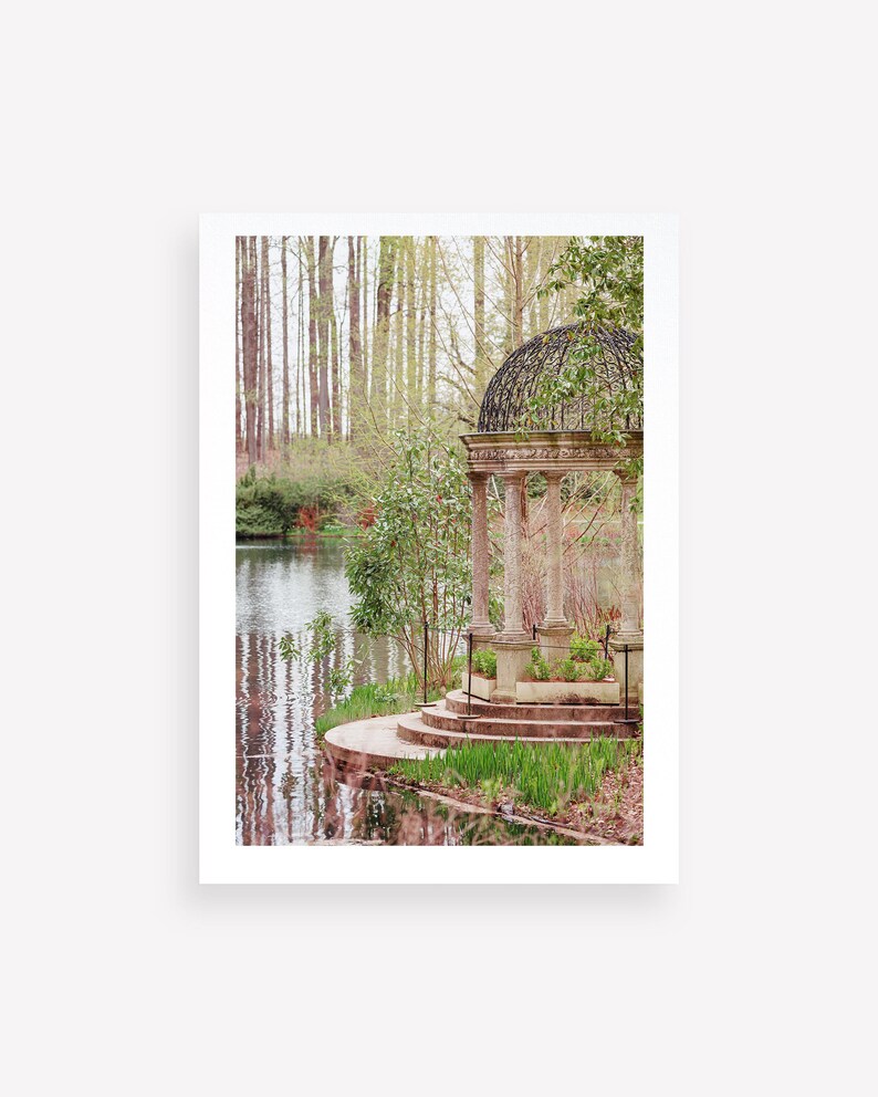 May include: A stone gazebo with a black metal roof sits on a small island in a pond. The gazebo has steps leading up to it and is surrounded by green trees and bushes.