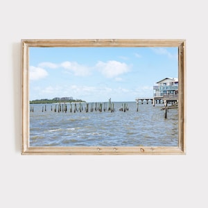 May include: A framed print of a coastal scene. The image features a body of water with wooden pilings, a building on a pier, and a distant island under a blue sky with clouds. The frame is made of light-colored wood.
