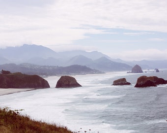 May include: A panoramic view of a rocky coastline with a hazy mountain range in the background. The ocean is a light blue with whitecaps and the rocks are a dark brown. The beach is a light brown with a few trees in the distance.