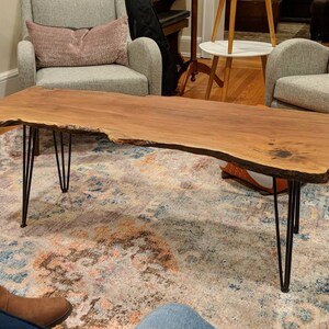 May include: A large, live edge wooden coffee table with a natural wood finish and black hairpin legs. The table is sitting on a patterned rug in a living room.
