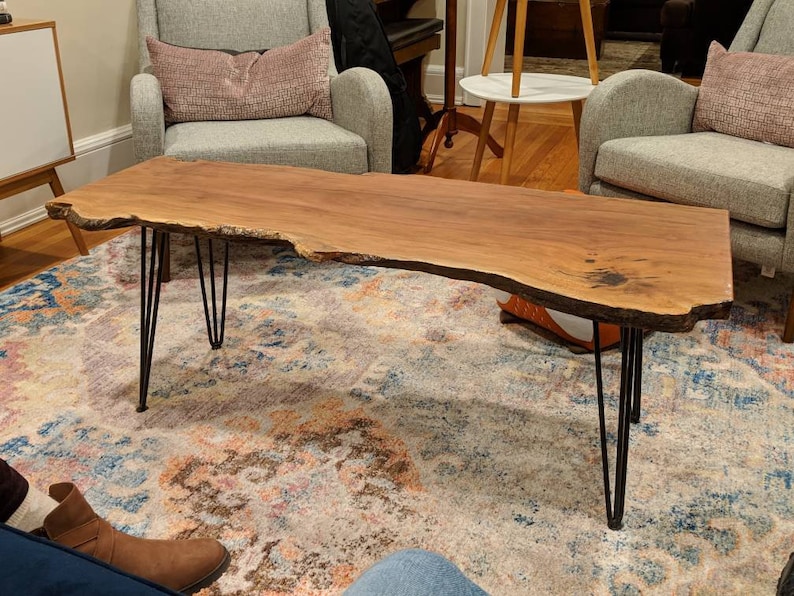 May include: A large, live edge wooden coffee table with a natural wood finish and black hairpin legs. The table is sitting on a patterned rug in a living room.