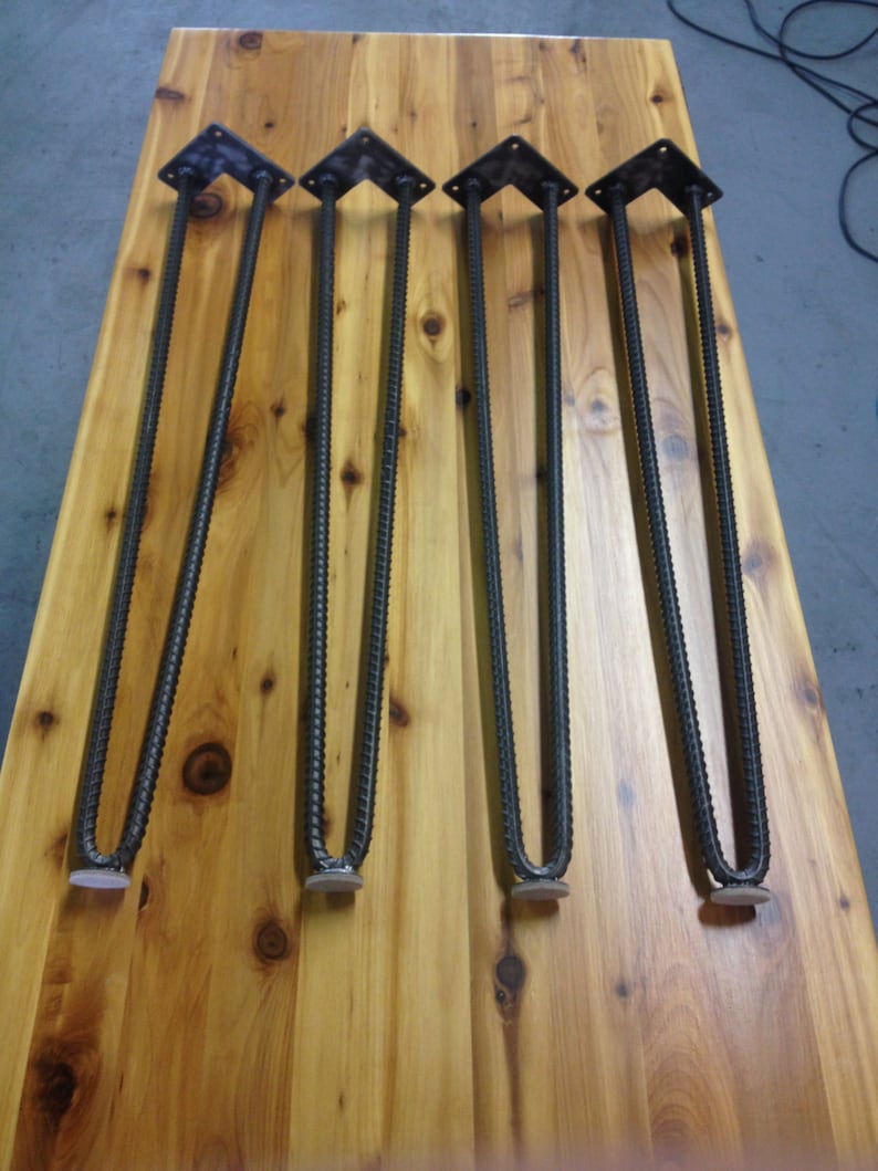 Puede incluir: Cuatro patas de horquilla de metal negro con almohadillas de fieltro blanco en la parte inferior. Las patas est&aacute;n dispuestas en una fila sobre una superficie de madera.