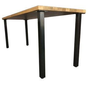 May include: A rectangular table featuring a light brown butcher block wood top with a visible grid pattern of wood grain segments. The table is supported by four straight, matte black metal legs attached with square mounting plates at each corner.