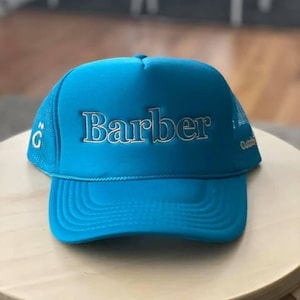 May include: A bright blue trucker hat with a white mesh back. The hat has the word "Barber" embroidered in white on the front.