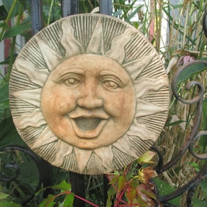 May include: A ceramic sun sculpture with a smiling face and a textured, white and brown finish. The sun is attached to a black metal fence.