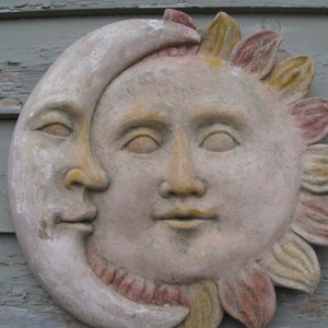 May include: A decorative wall sculpture of the sun and moon, with the sun's face in the centre and the moon's crescent shape surrounding it. The sun has a yellow and orange face with a pink and yellow flower-like design around it. The moon is a light grey colour.