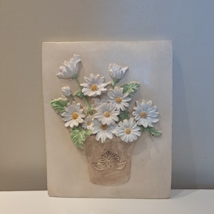 May include: A white ceramic wall plaque featuring a 3D relief design of a bouquet of white daisies in a brown pot.
