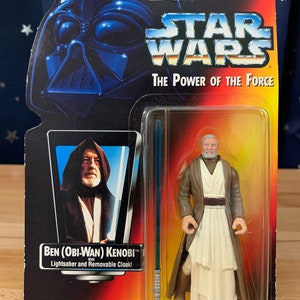 Op de afbeelding: Een Star Wars-actiefiguur van Obi-Wan Kenobi, die een bruine robe en een witte tuniek draagt en een blauw lichtzwaard vasthoudt. De figuur is verpakt in een plastic blisterverpakking met een gele en rode achtergrond. De verpakking toont het Star Wars-logo en de tekst "The Power of the Force".