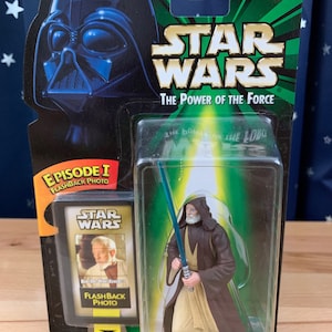 Op de afbeelding: Een Star Wars-actiefiguur van Ben (Obi-Wan) Kenobi met een blauw lichtzwaard. De figuur is verpakt in een plastic blisterverpakking met een groene en zwarte achtergrond. De verpakking heeft het Star Wars-logo en de woorden "The Power of the Force".