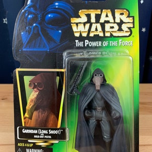 Op de afbeelding: Een Star Wars-actiefiguur van Garindan, ook bekend als Long Snoot, uit de Power of the Force-collectie. De figuur draagt een zwarte cape en een gasmasker. De figuur houdt een blaster in zijn rechterhand. De figuur is verpakt in een plastic blisterverpakking met een groene achtergrond. De verpakking bevat de tekst "Garindan (Long Snoot)™ with Hold-Out Pistol" en "Ages 4 & Up".