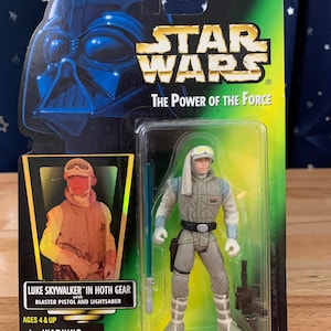 Op de afbeelding: Een Star Wars-actionfiguur van Luke Skywalker in zijn Hoth-uitrusting. De figuur draagt een wit en grijs sneeuwpak met een witte sjaal en een bruine helm. Hij houdt een blauwe lightsaber en een zwart blasterpistool vast. De verpakking is groen en heeft het Star Wars-logo en de woorden "The Power of the Force".