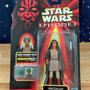 Puede incluir: Una figura de acción de Star Wars Episodio I de Adi Gallia con un sable de luz rojo. La figura está empaquetada en un blíster de plástico con un fondo rojo, negro y dorado. El empaque presenta el logotipo de Star Wars y el texto "Now figures talk!" con un chip CommTech.