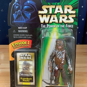 Op de afbeelding: Star Wars The Power of the Force actionfiguur van Chewbacca in een besneeuwde omgeving met een bowcaster-geweer. De figuur is verpakt in een groene en zwarte doos met een flashbackfoto van Chewbacca.