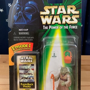 Op de afbeelding: Een Star Wars Episode I Flashback Photo actionfiguur van Yoda met een stok en een kokende pot. De figuur is verpakt in een groene en witte doos met een afbeelding van Yoda op de voorkant.