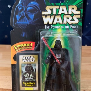 Op de afbeelding: Een Star Wars-actiefiguur van Darth Vader met een rood lichtzwaard. De figuur is verpakt in een plastic blisterverpakking met een groene en zwarte achtergrond. De verpakking heeft het Star Wars-logo en de woorden "The Power of the Force".