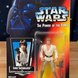 Op de afbeelding: Een Star Wars action figure van Luke Skywalker uit de The Power of the Force serie. De figuur draagt een witte tuniek en een bruine broek. Hij houdt een blaster en een lightsaber vast. De verpakking is rood, geel en zwart met het Star Wars logo.