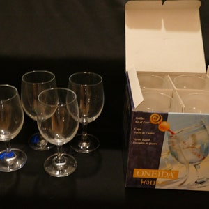 May include: A set of four clear wine glasses with a box. The glasses are on a black surface. The box is white and purple with the words "Goblet Set of Four" and "ONEIDA Holiday" printed on it.