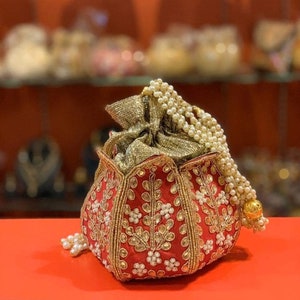 May include: A red and gold embroidered clutch bag with a pearl and gold chain strap. The bag is decorated with floral and leaf patterns.